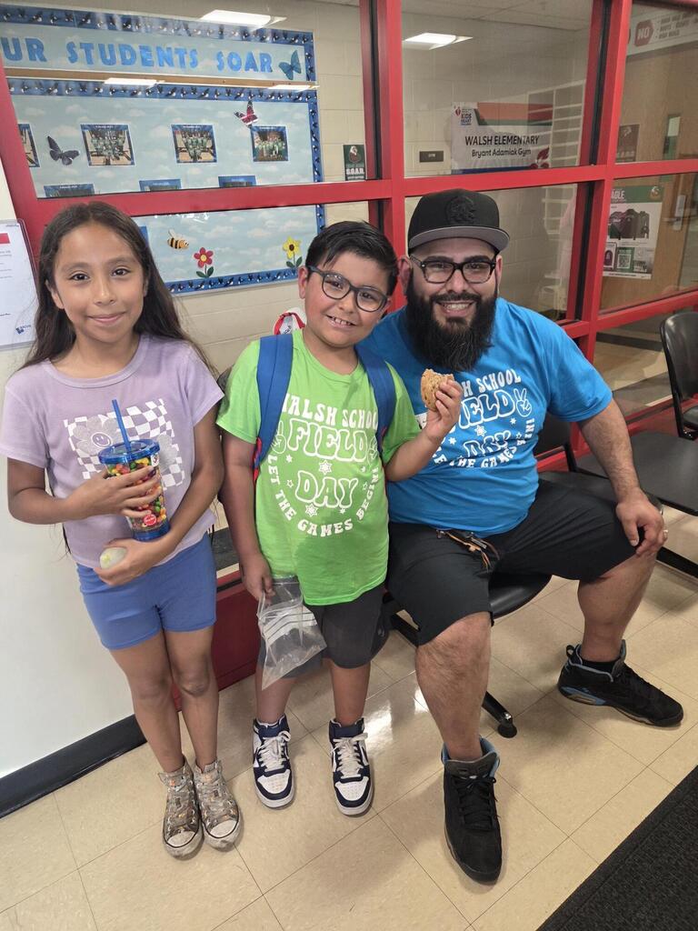 A man in a blue t-shirt and baseball cap smiles while holding a cookie, standing next to two smiling children in front of a school bulletin board.