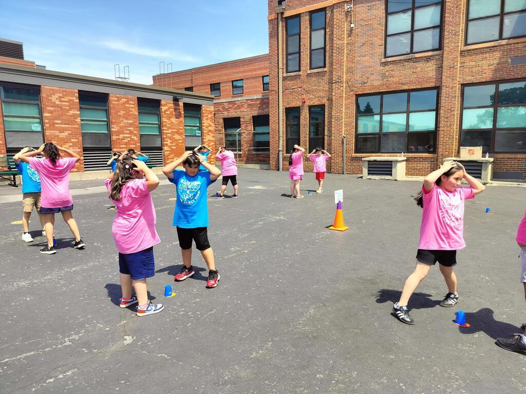 Children in pink and blue shirts play a game in a schoolyard, balancing cones on their heads. They're smiling under a clear blue sky, exuding joy.