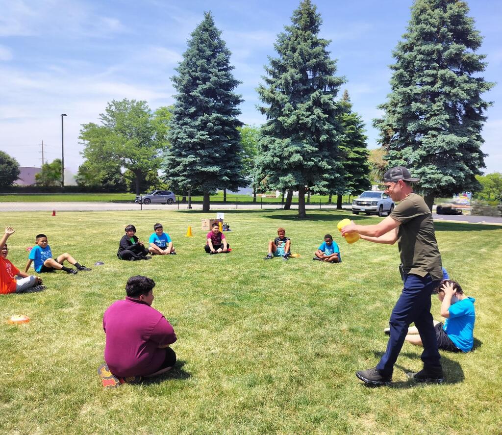 A man in a cap carries a yellow sponge around children sitting in circle on a grassy lawn near tall pine trees.