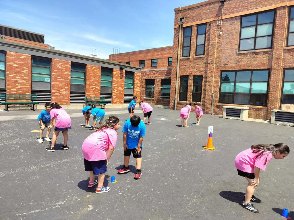 Children in blue and pink shirts participate in an outdoor activity in front of a brick school building.