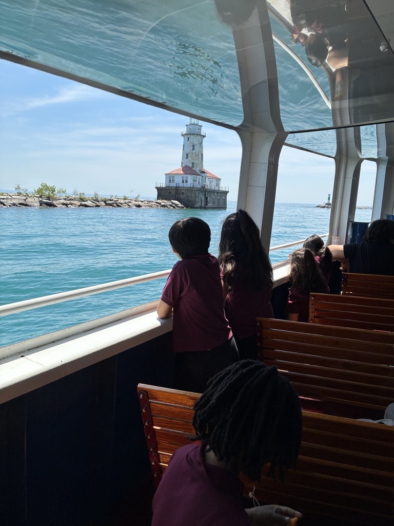 Children in maroon shirts look out from a boat at a distant lighthouse on a sunny day. The turquoise water and light-filled cabin create a serene vibe.