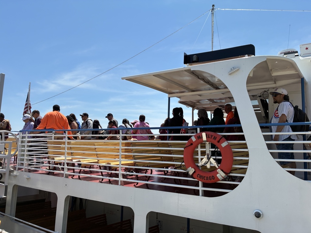 A group of people stands on a ferry deck under a clear blue sky, some seated on yellow benches. A red lifebuoy reads "Marilyn Chicago." The atmosphere is sunny and relaxed.