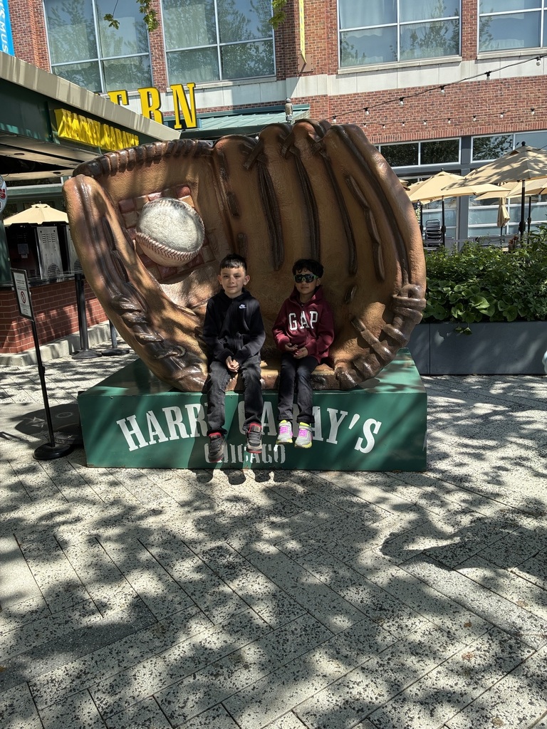 Two children sit on a giant baseball glove sculpture outside Harry Caray's restaurant in Chicago. It's a sunny day with dappled shadows on the ground.