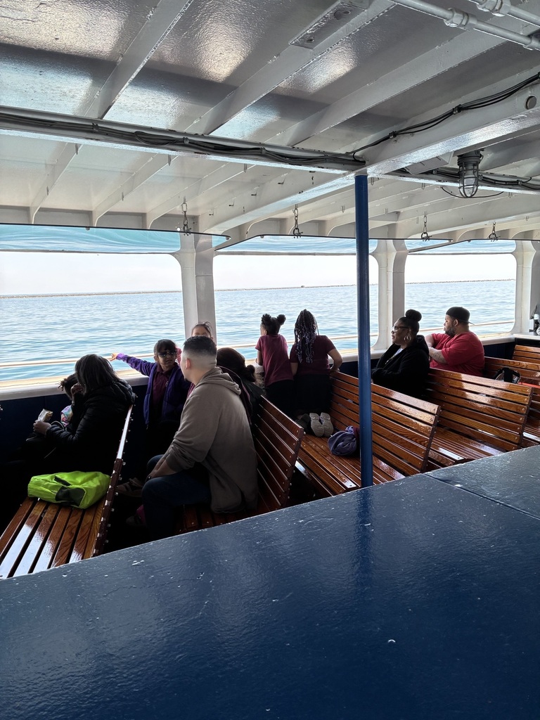 People are seated on wooden benches inside a ferry, looking out at the calm sea. The mood is relaxed, and sunlight filters through the canopy above.