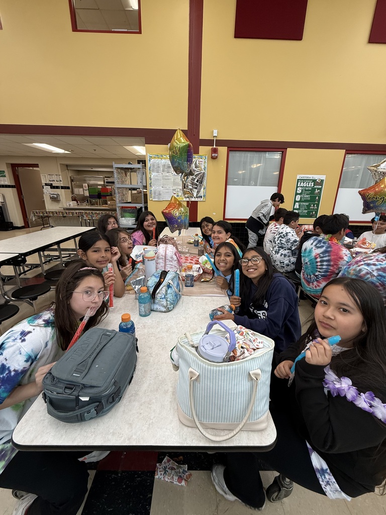 A group of smiling kids in a cafeteria enjoy snacks at a long table, surrounded by colorful bags and balloons. The atmosphere is lively and joyful.