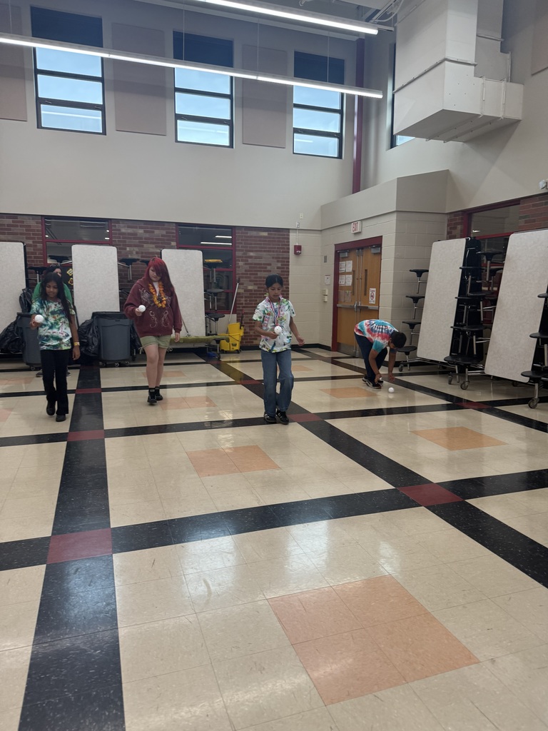 Students in tie-dye shirts play with small balls in a large room with tiled floors and high windows. The atmosphere is casual and playful.