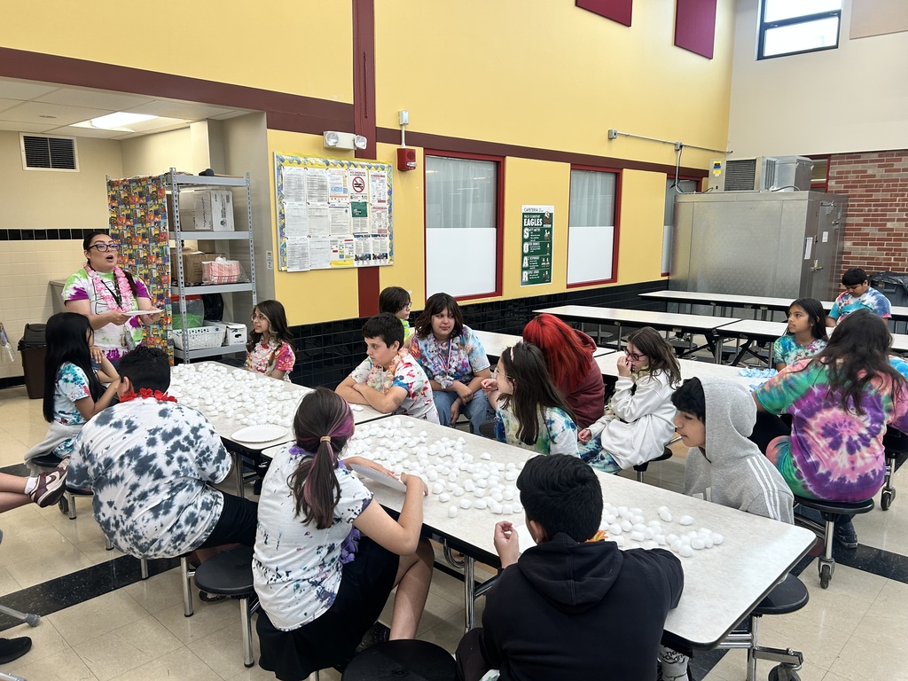 Students in tie-dye shirts sit around cafeteria tables covered with small white items, listening to a woman standing and speaking energetically.