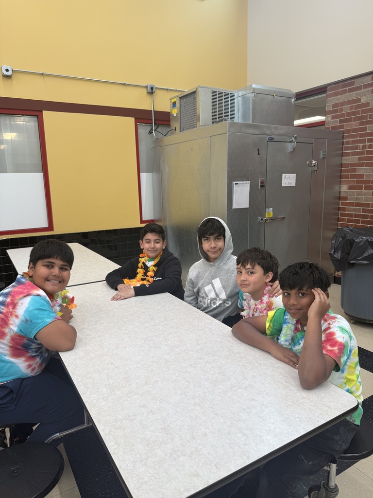 Five boys sit at a cafeteria table, wearing tie-dye shirts and leis, smiling. The room has yellow walls and a metal storage unit. A joyful, casual atmosphere.