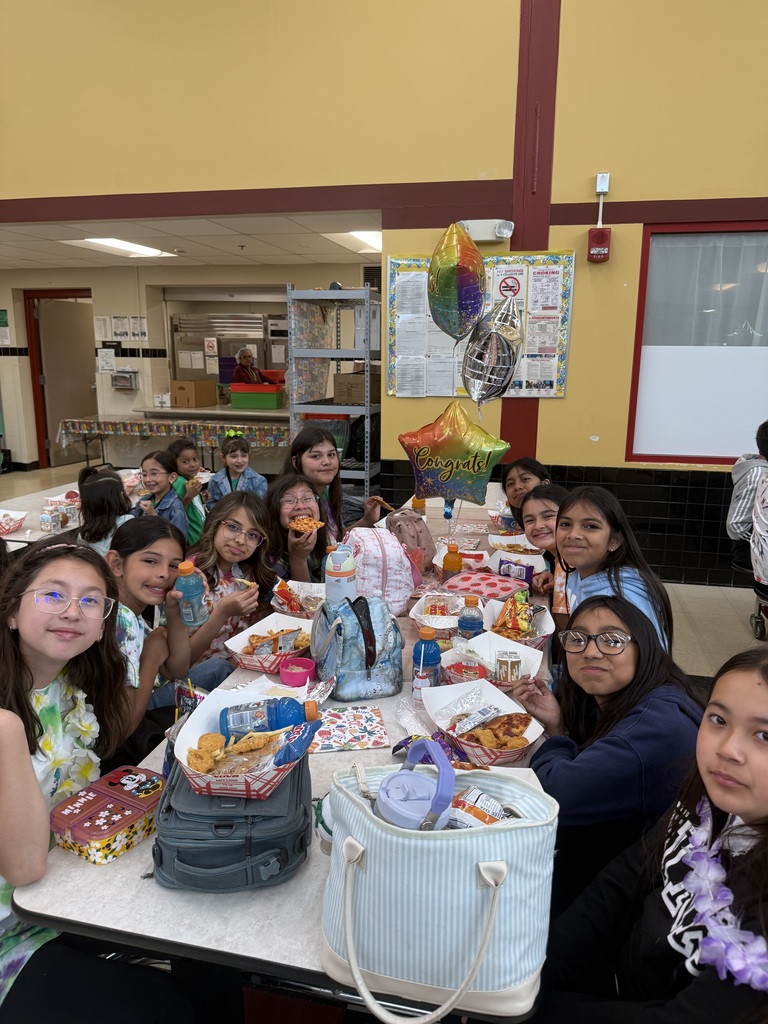 A group of young students smiling and eating lunch at a school cafeteria table. Festive "Congrats!" balloons enhance the celebratory mood.