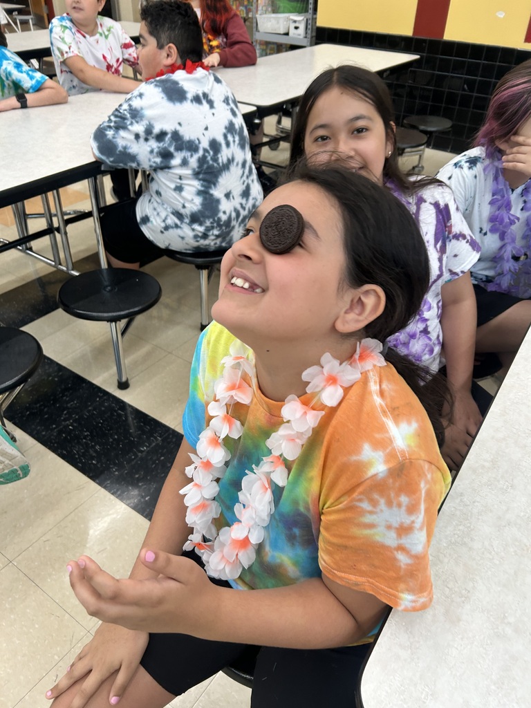 A smiling child with an Oreo cookie on their forehead, wearing a colorful tie-dye shirt and flower lei, sits in a lively cafeteria setting.