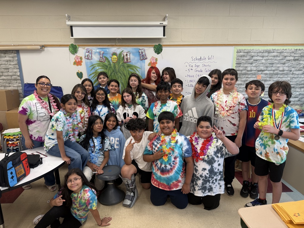 A group of children and one adult in tie-dye shirts and leis posing happily in a classroom. The background features a tropical-themed decoration.