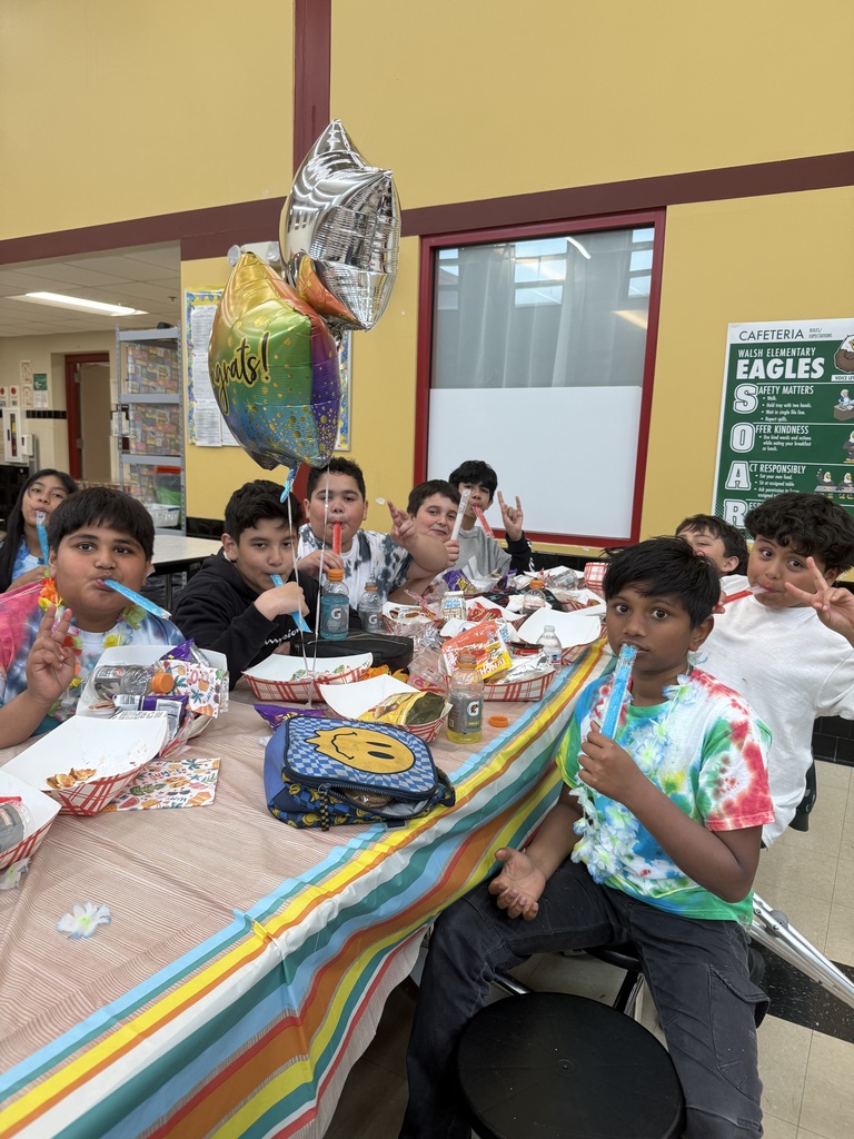 A group of kids at a decorated table enjoying snacks and drinks. They're wearing colorful shirts and holding popsicles, with balloons reading "Congrats!" in the background.