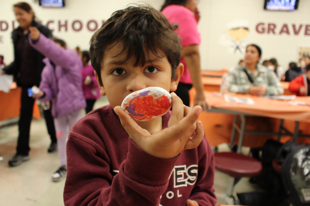 A young child in a sweatshirt holds up a painted rock, displaying it proudly. The background shows a school cafeteria with people seated and walking around.