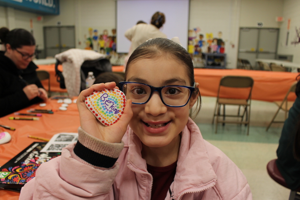 A young girl with glasses holds a decorated stone reading "Be Kind," smiling in a room with people engaged in crafts at an art event.