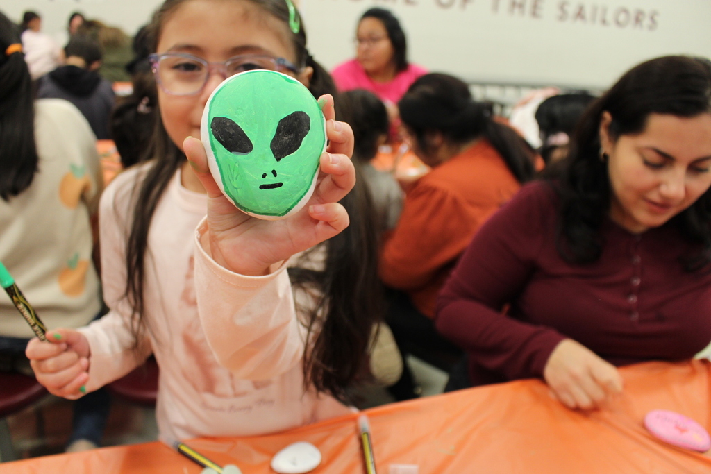 A young girl with glasses holds up a painted stone featuring a green alien face. She's surrounded by others painting at a table. The scene is lively and creative.