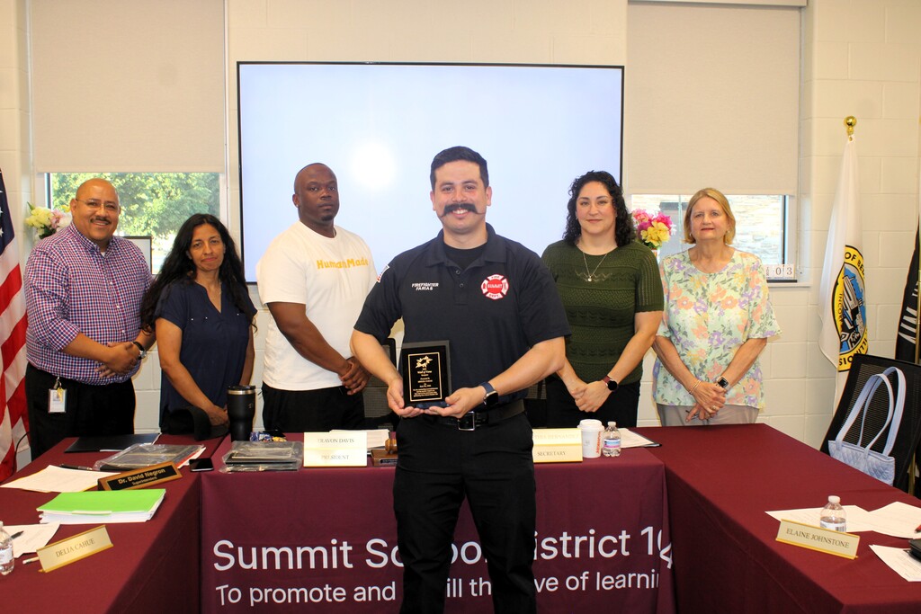 A man stands smiling, holding a plaque in a meeting room with five colleagues behind a table. The setting is positive and professional.