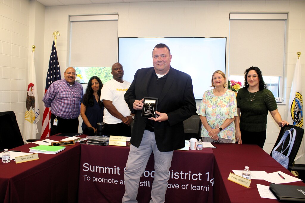 A group of five people smiles in a meeting room. One person holds a plaque. They're standing behind a table with Summit School District 104 signs.