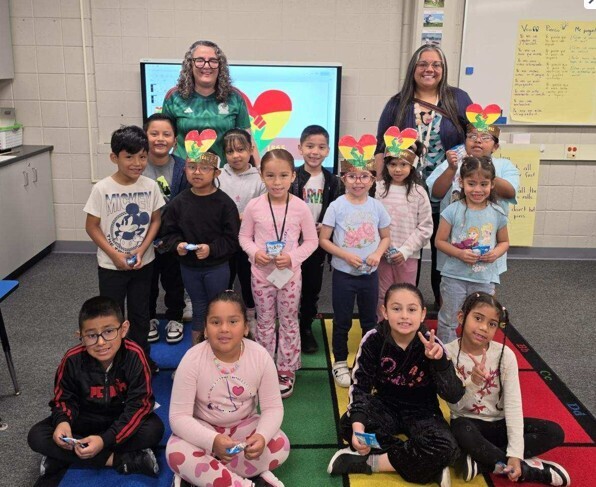 A group of children and two adults smile in a classroom. Some kids wear heart-shaped headbands. The mood is joyful and the setting is colorful.