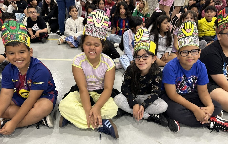 Children in colorful, handmade crowns sit cross-legged on the floor, smiling. Their crowns feature "Juneteenth" with hand designs. The mood is joyful and celebratory.