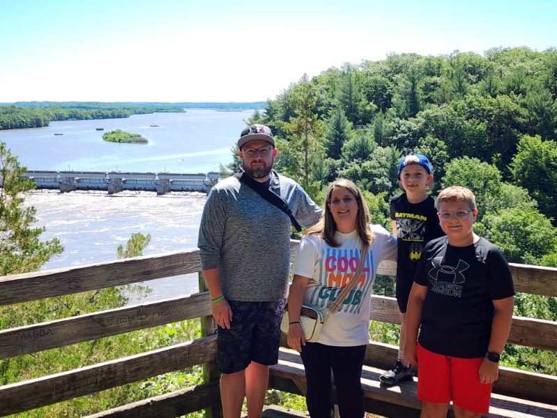 A family of four poses on a wooden deck overlooking a scenic river and lush forest. They smile warmly, enjoying a bright, sunny day outdoors.