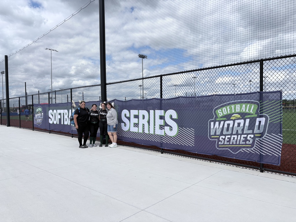 Four people stand smiling in front of a fence with a "Softball World Series" banner, under a cloudy sky on a sports field.