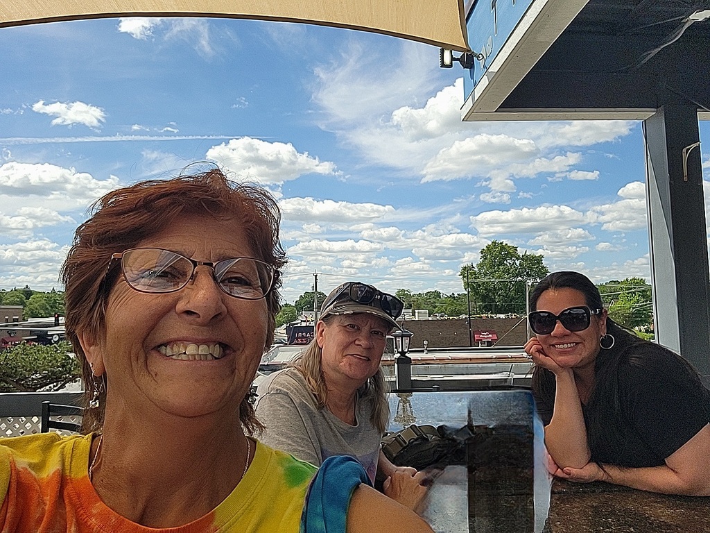 Three people enjoy a sunny day on a patio. The foreground shows a smiling individual in a tie-dye shirt. Two others sit at a table with a blue sky and clouds above.