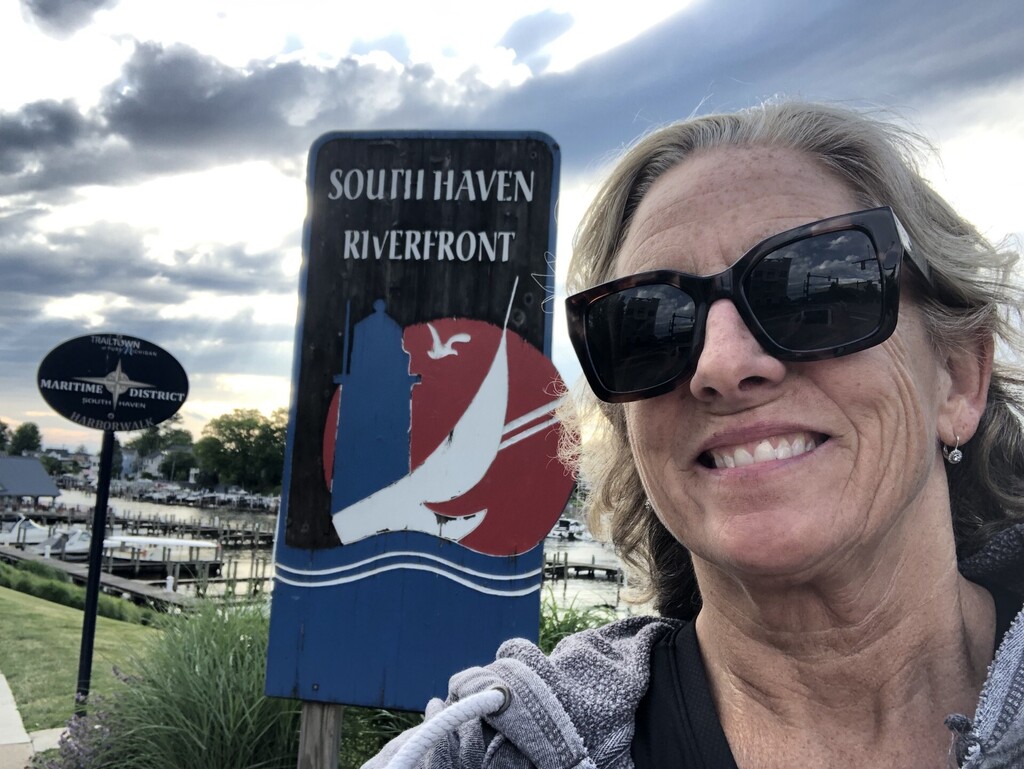 Smiling woman in sunglasses takes a selfie at South Haven Riverfront. A sign and cloudy sky are visible with a marina in the background.