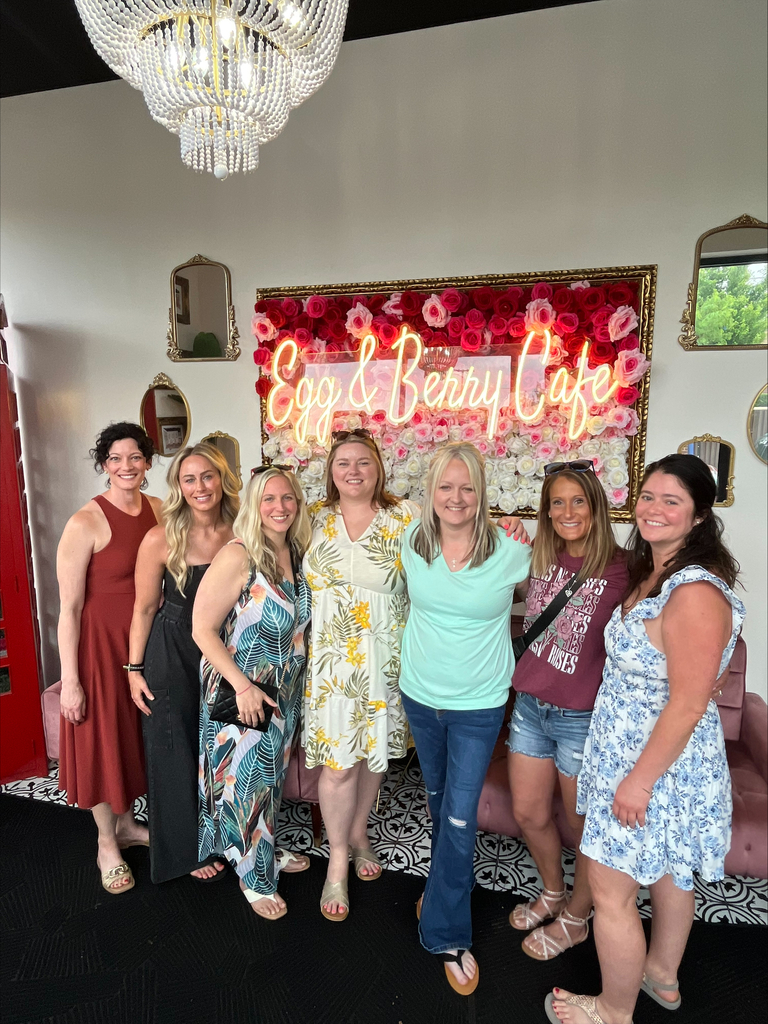 A group of seven women smiling, standing inside "Egg & Berry Cafe." Behind them is a wall with a floral design and neon sign. Elegant chandelier overhead.