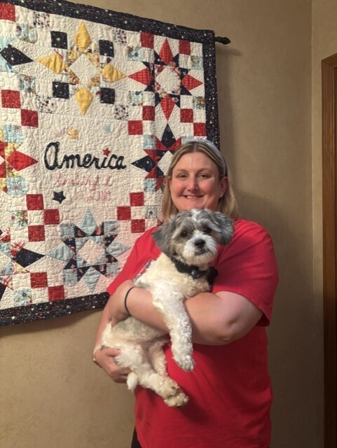 A woman in a red shirt smiles while holding a small, fluffy dog. Behind them is a quilt with a star pattern, featuring the word "America."