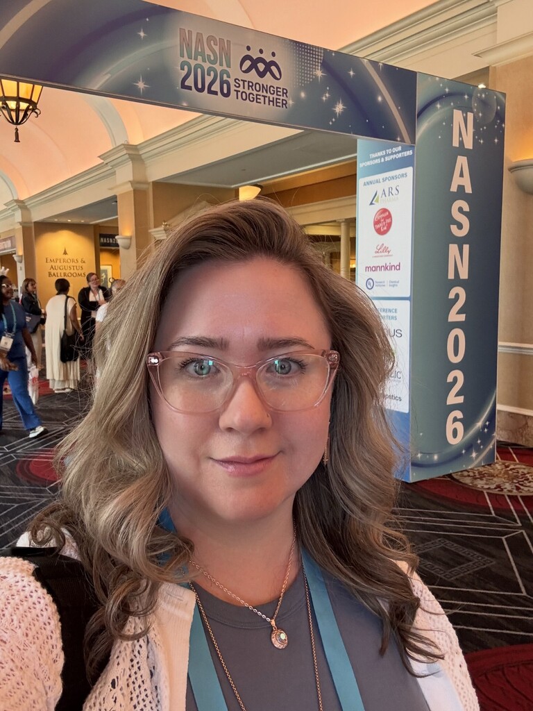 Woman with glasses and long hair smiles at a conference. Background shows a banner for "NASN 2026 Stronger Together" and a list of sponsors.
