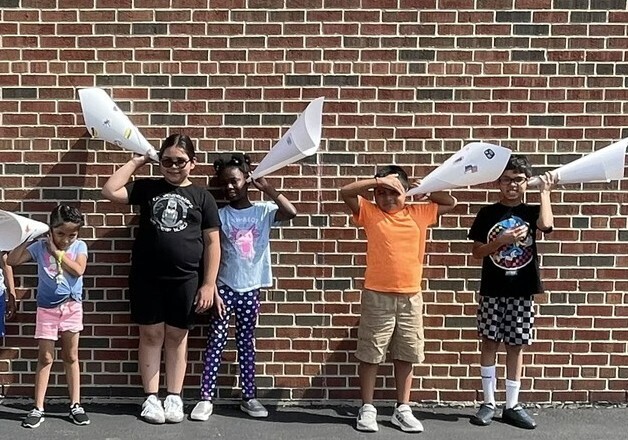 Six children stand against a brick wall, each holding large paper cones like megaphones. They appear playful and joyful on a sunny day.
