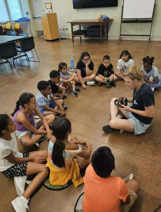 A group of children sit in a circle on the floor, attentively listening to a woman holding a small animal. The room is bright and educational, conveying curiosity.