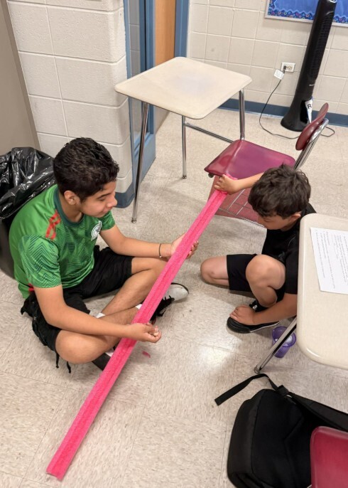 Two boys are seated on a classroom floor, stretching a long pink streamer between them. The environment is casual and collaborative, with desks around.