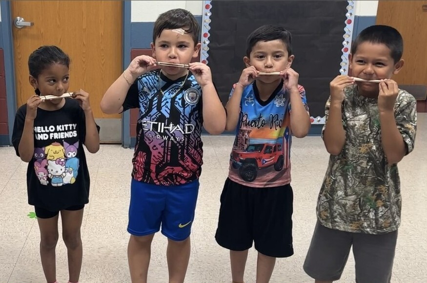 Four children stand in a classroom, each holding a wooden stick up to their mouths as pretend mustaches. They are smiling and appear playful.