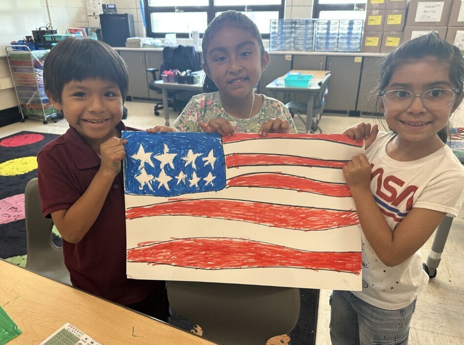 Three smiling children in a classroom hold a handmade American flag drawing with red stripes and white stars on a blue background, conveying pride and creativity.