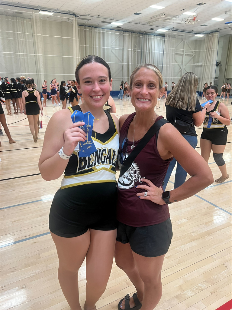 Cheerleader in a black and gold "Bengals" uniform holds blue ribbons, smiling with a woman in a maroon tank top on a gym floor; a joyful atmosphere.