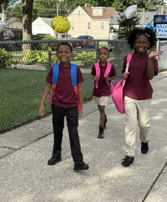 Three children walk on a sidewalk, wearing maroon school uniforms and smiling. Two carry pink backpacks, and one has a blue backpack. Balloons float nearby.