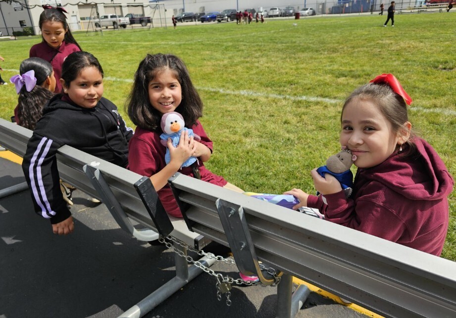 . Three girls sit on a bench, happily playing with their toys in a sunny outdoor setting.
