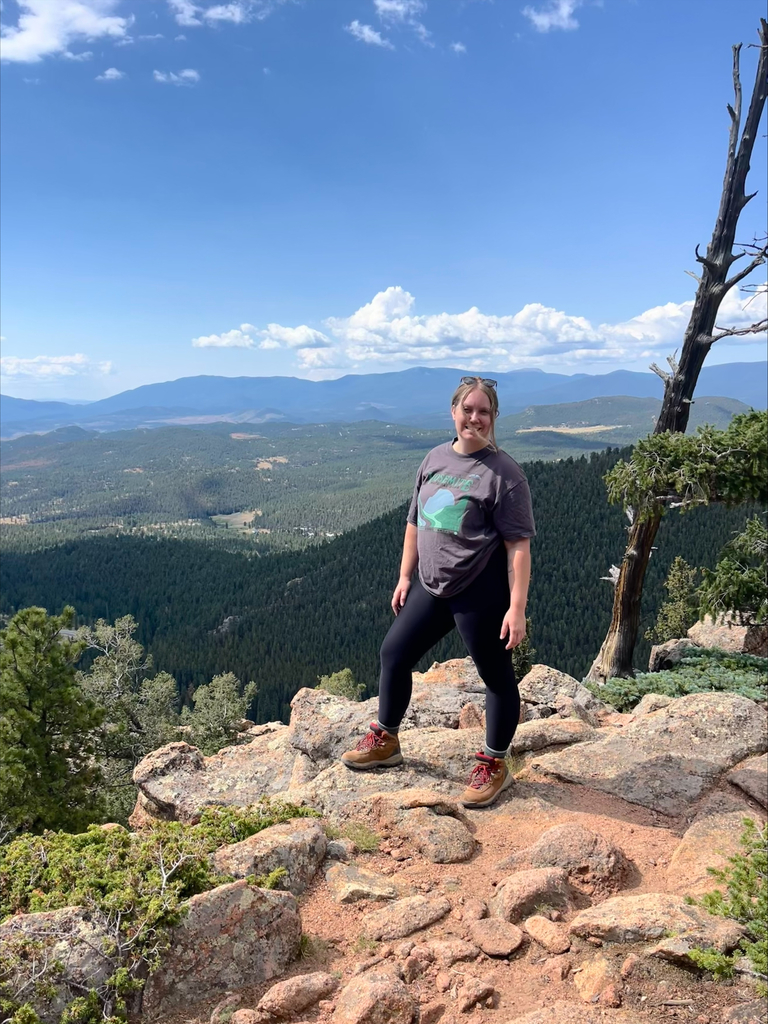 A person smiles atop a rocky mountain viewpoint, surrounded by lush greenery and expansive views of distant hills under a bright blue sky with fluffy clouds.