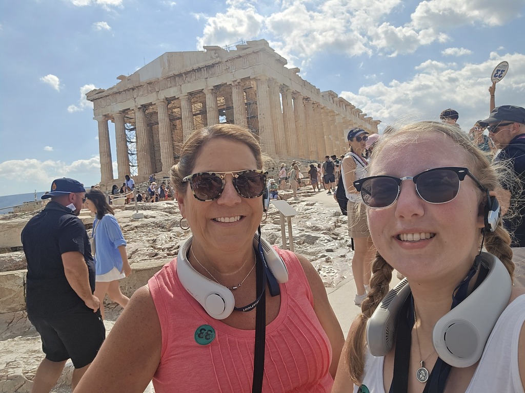 Two women wearing headphones and sunglasses pose in front of the Parthenon in Athens on a sunny day, with other tourists and the ancient ruins visible in the background.
