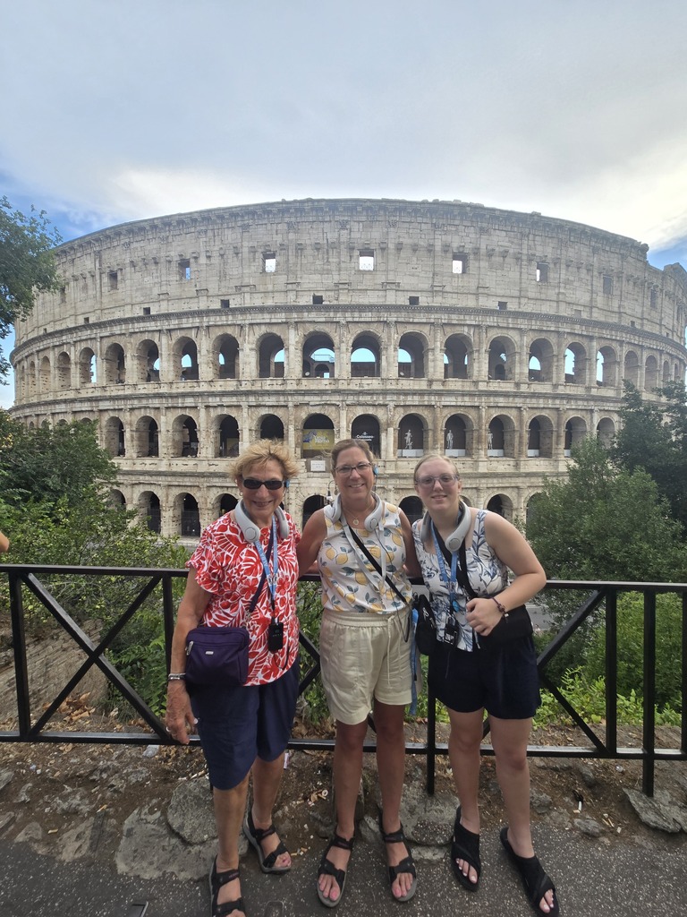 Three women smile for a photo in front of the Colosseum, showcasing the iconic ancient structure in the background.
