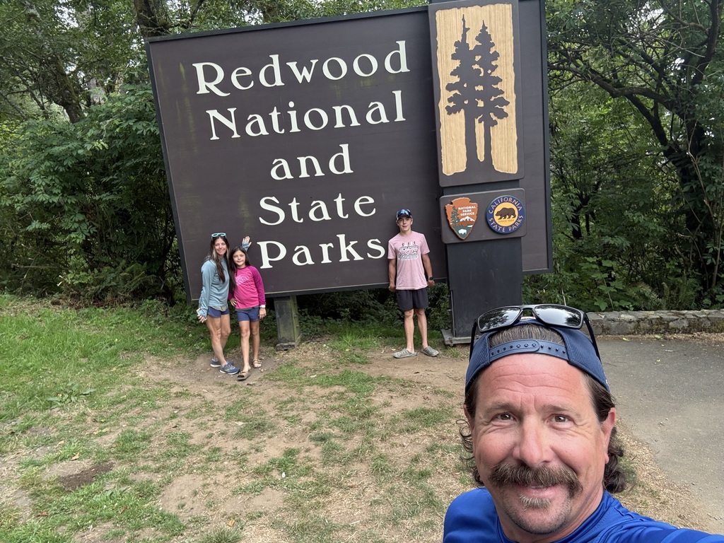 A family selfie featuring a man with a woman and two children in front of the Redwood National and State Parks sign.