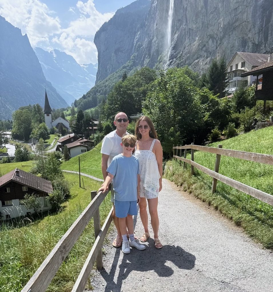 A family stands on a sunlit path with a scenic backdrop of lush green hills, a tall waterfall, and a quaint chapel. The mood is serene and joyful.