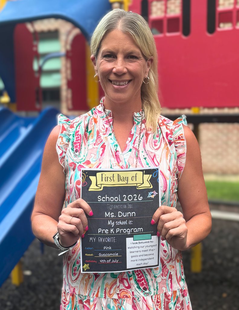A woman holding a "First Day of School 2026" sign. She is wearing a colorful dress and standing in front of a playground.