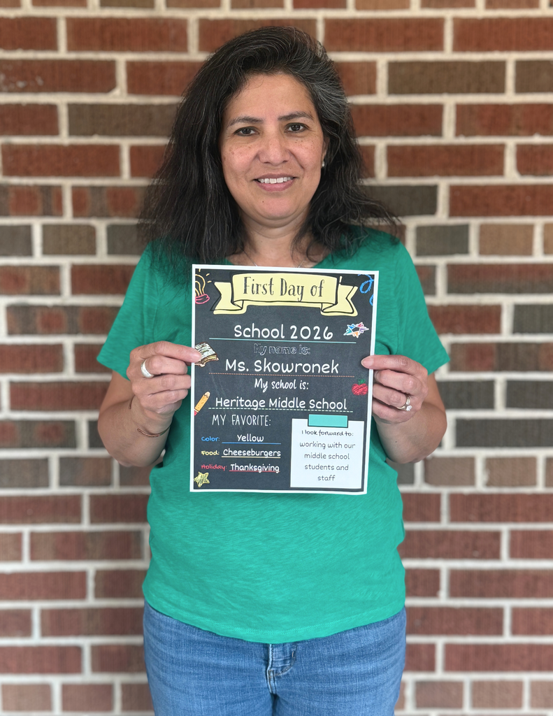A woman holding a sign that says "First Day of School 2026" with her name and school information on it, standing in front of a brick wall.