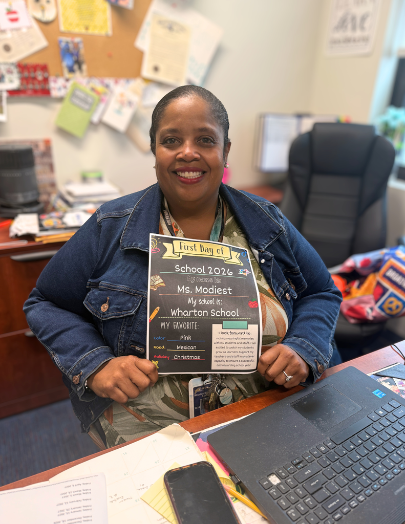 A smiling woman sitting at a desk holds a sign that reads "First Day of School 2022, Ms. Modiest, Wharton School." The desk has items like a laptop, papers, and personal decorations.