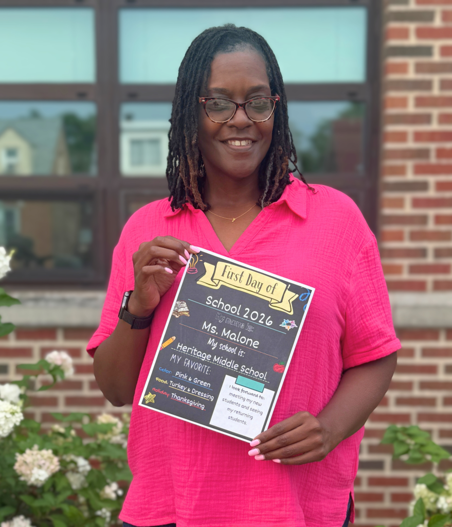 A woman in a bright pink shirt holds a sign that says "First Day of School 2026" and includes her name, "Ms. Malone," and "Heritage Middle School."