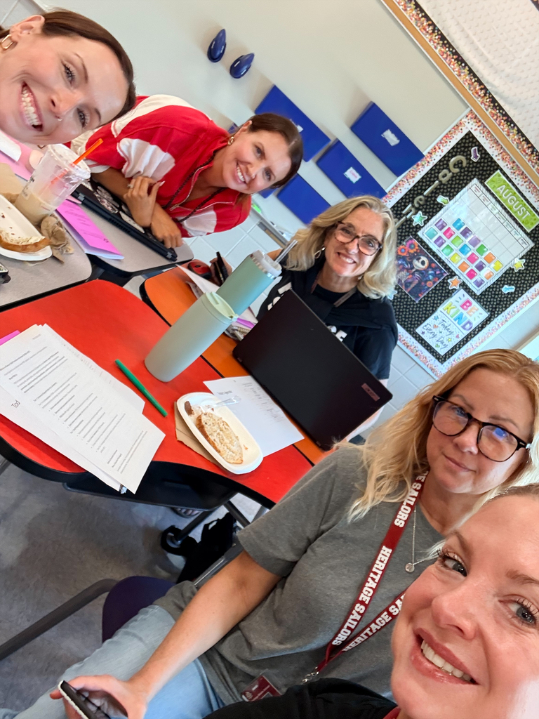 Five smiling women pose for a selfie in a colorful classroom, with a laptop, papers, lunch plates, and a vibrant bulletin board behind them.
