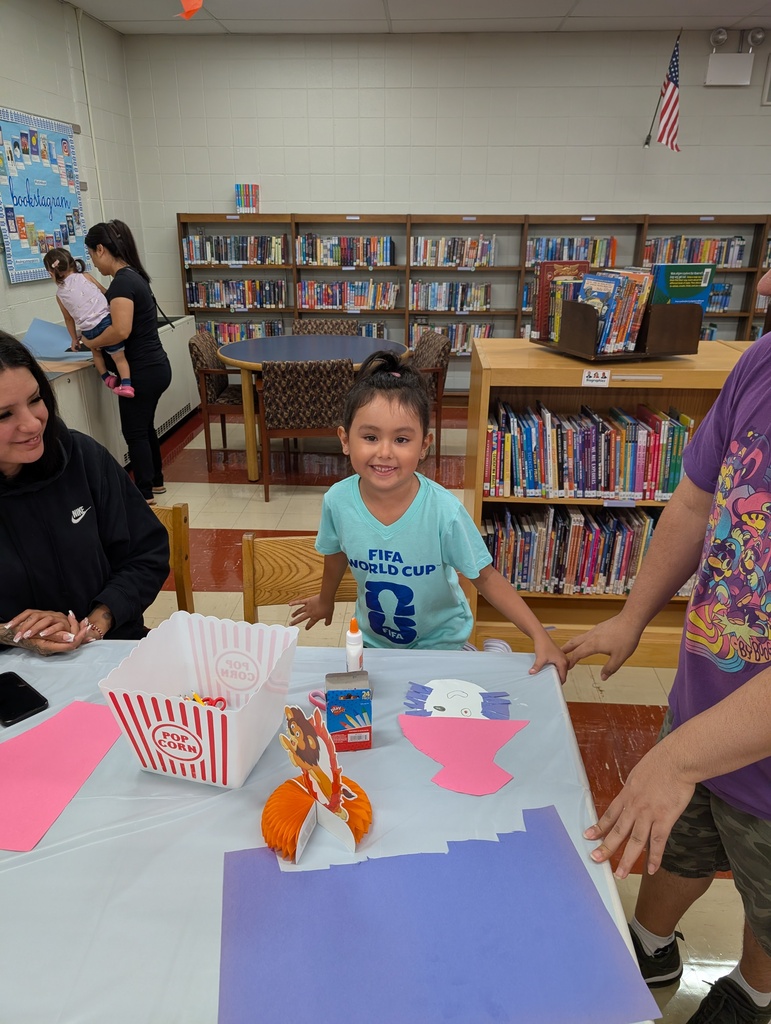 Smiling child creates a colorful paper craft at a library table, with adults nearby, bookshelves behind, and a popcorn box in view.