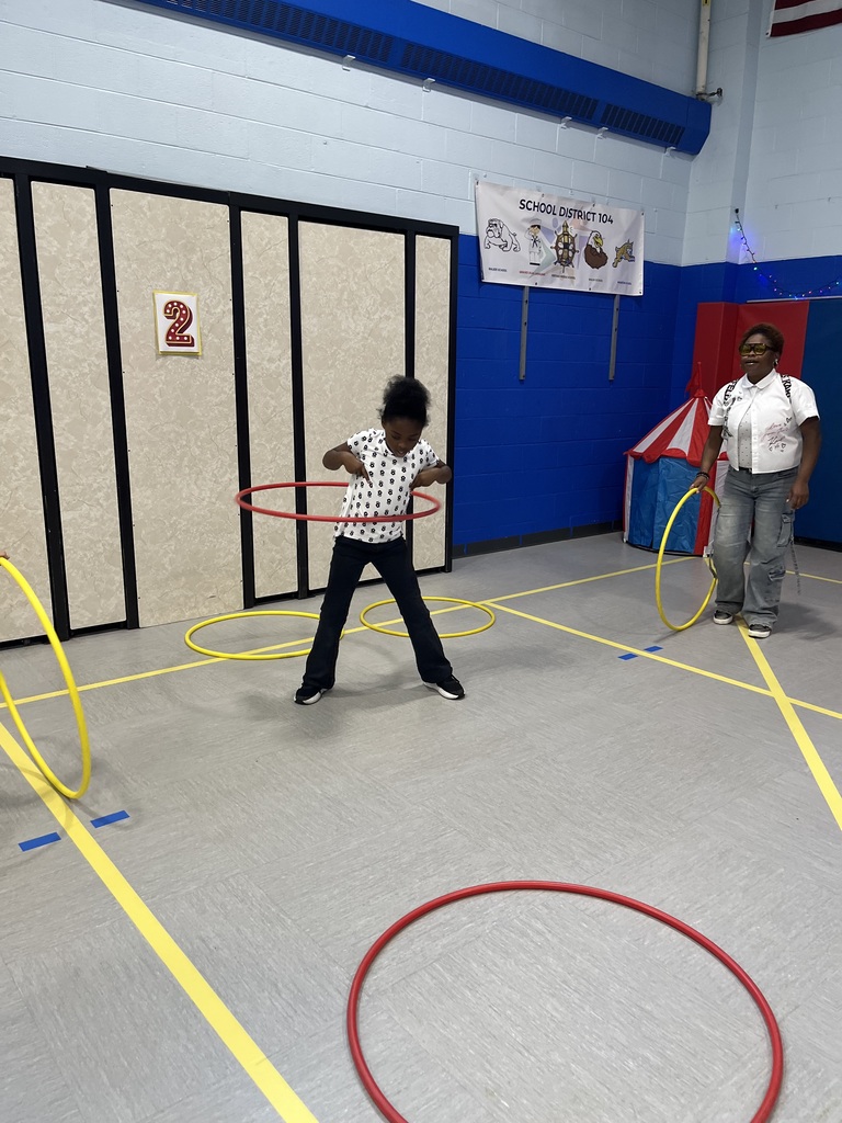Child practices with a red hula hoop in a colorful school gym while an adult supervises; scattered hoops nearby create a playful tone.